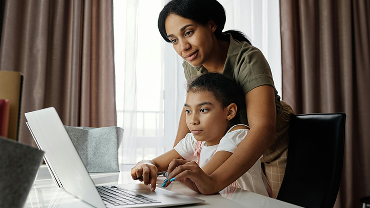 Child working on laptop at desk with mother behind her.