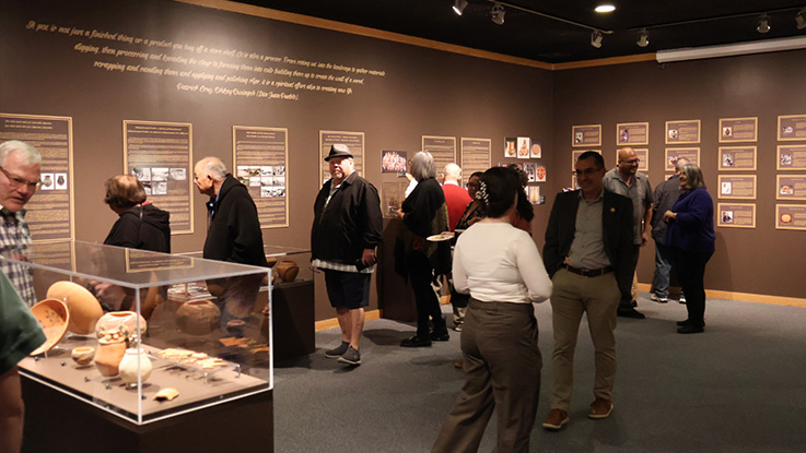 People standing in exhibit with pottery under glass in middle of room.